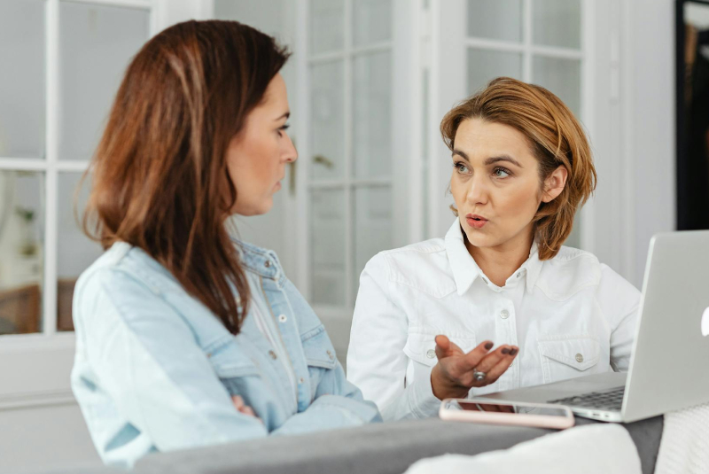 Zwei Frauen im Gespräch an einem Tisch, eine erklärt etwas vor ihrem Laptop