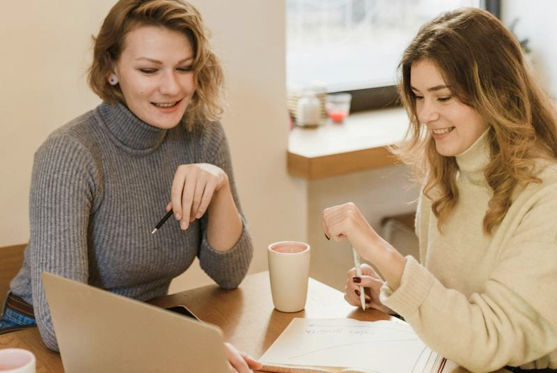Zwei lächelnde Frauen arbeiten gemeinsam mit Laptop und Notizbuch an einem Tisch